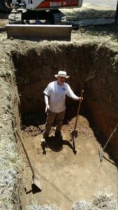 Mini excavator digging hole for a septic tank, man leveling by hand with shovel