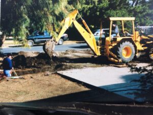 Backhoe digging trench by cement slab next to road in neighborhood