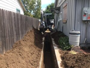 Side yard of house site work with man in trench hand digging, mini excavator in background
